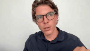 Primer plano de Oriol Blanch de Bitvavo, un hombre joven con gafas de carey y camisa polo azul oscuro, hablando y mirando hacia la izquierda del encuadre contra un fondo de pared blanca plana.