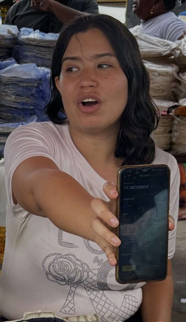 Un plano medio de una mujer joven con cabello oscuro y una franela blanca estampada, de pie detrás de lo que parece un puesto de mercado al aire libre. Ella extiende su brazo derecho hacia adelante, sosteniendo un teléfono inteligente con la pantalla hacia la cámara. La pantalla del teléfono muestra una interfaz de aplicación financiera.