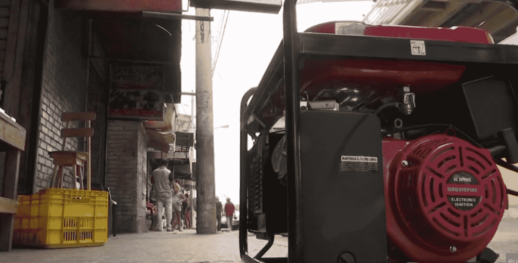 Se observa una planta eléctrica portátil de color roja, ubicada en el suelo a la derecha. El fondo muestra varias personas caminando por la calle.