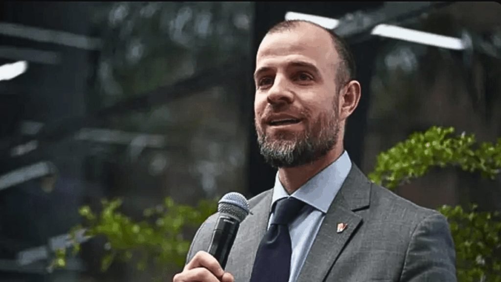 Fotografía de medio cuerpo de Román Maniglia, vestido con traje gris, camisa azul claro y corbata oscura, sosteniendo un micrófono negro con la mano derecha mientras habla en un evento público con vegetación de fondo.