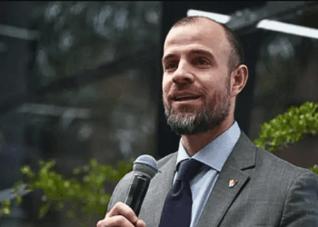 Fotografía de medio cuerpo de Román Maniglia, vestido con traje gris, camisa azul claro y corbata oscura, sosteniendo un micrófono negro con la mano derecha mientras habla en un evento público con vegetación de fondo.