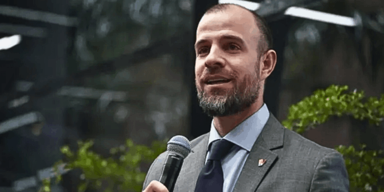 Fotografía de medio cuerpo de Román Maniglia, vestido con traje gris, camisa azul claro y corbata oscura, sosteniendo un micrófono negro con la mano derecha mientras habla en un evento público con vegetación de fondo.