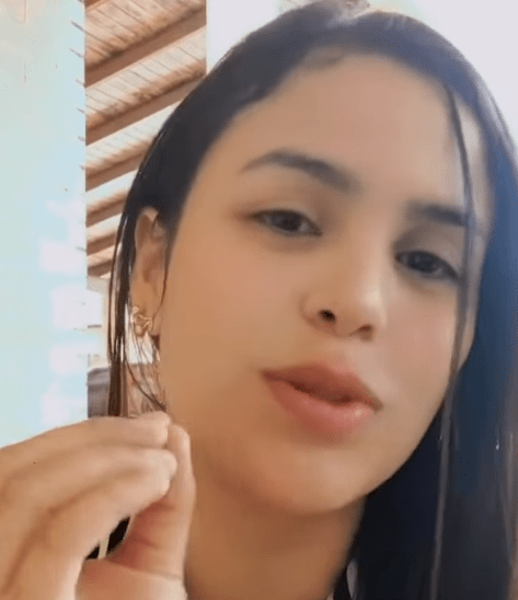 Primer plano de Yossem Moreno, una mujer joven con cabello castaño oscuro, mientras habla. Su mano derecha está levantada con los dedos juntos cerca de su boca en un gesto de explicación o énfasis. Viste un top blanco y un pequeño pendiente de oro es visible en su oreja. El fondo es un área exterior borrosa pero brillantemente iluminada con vigas de madera de un techo. La imagen es un fotograma de video.