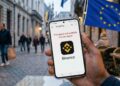 Mano sosteniendo un móvil en una terraza europea mostrando en pantalla el logo de Binance y un mensaje de restricción.