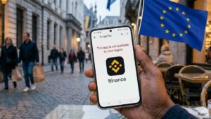 Mano sosteniendo un móvil en una terraza europea mostrando en pantalla el logo de Binance y un mensaje de restricción.