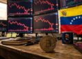 Bitcoin sobre una mesa con gráficos en rojo en el fondo y la bandera de Venezuela.