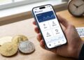 Mano sosteniendo un teléfono móvil con la aplicación BlueWallet abierta en una mesa junto a monedas de bitcoin.