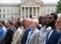 Un grupo de personas aparecen en plano medio mirando hacia el frente y un edificio institucional se observa en el fondo.