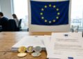 Criptomonedas en una mesa con un documento que dice Ley MiCA y la bandera de la UE en el fondo