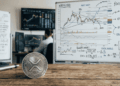 Primer plano: moneda física de XRP. Fondo: pizarra con análisis de precio.