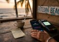 Mano femenina sosteniendo un teléfono inteligente sobre un mostrador de madera en un café costero con vista al Pacífico. Pantalla de billetera Bitcoin muestra "cero transacciones en el último mes". Al lado, datáfono bancario encendido. Taza vacía, recibo arrugado, papel con el número "31". Letrero desgastado de "Bitcoin Beach" en la pared. Luz cálida de atardecer. Estilo cinematográfico.