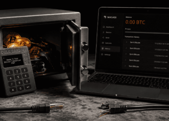 Fotografía conceptual en tono oscuro que muestra una billetera física Coldcard MK4 apoyada sobre una caja fuerte de metal abierta llena de monedas de Bitcoin. A la derecha, la pantalla de una computadora portátil en modo oscuro muestra la aplicación Wasabi Wallet con un balance en cero y varias salidas de fondos recientes. En primer plano hay un cable de red cortado sobre la superficie de piedra.