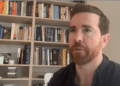 Un plano medio de James O'Beirne, un hombre con cabello castaño corto y una barba roja bien cuidada, que lleva una camisa azul oscura abotonada. Está mirando ligeramente hacia la izquierda de la cámara con una expresión de concentración mientras habla. Está sentado frente a una estantería de madera bien llena de libros con una pared gris claro a su derecha. La iluminación es suave y uniforme.