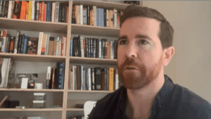 Un plano medio de James O'Beirne, un hombre con cabello castaño corto y una barba roja bien cuidada, que lleva una camisa azul oscura abotonada. Está mirando ligeramente hacia la izquierda de la cámara con una expresión de concentración mientras habla. Está sentado frente a una estantería de madera bien llena de libros con una pared gris claro a su derecha. La iluminación es suave y uniforme.