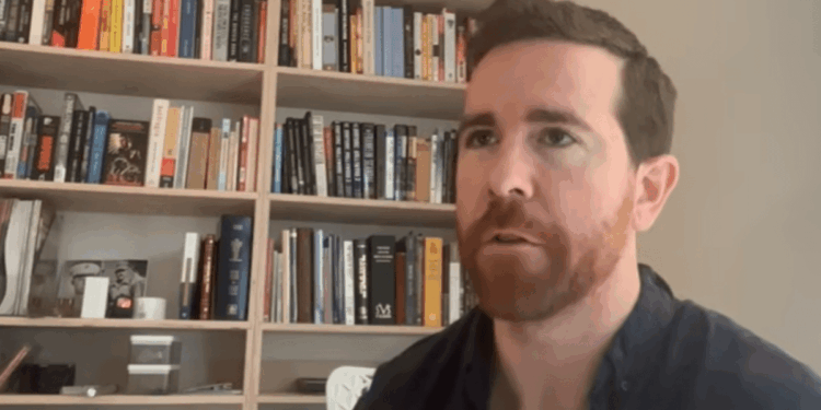 Un plano medio de James O'Beirne, un hombre con cabello castaño corto y una barba roja bien cuidada, que lleva una camisa azul oscura abotonada. Está mirando ligeramente hacia la izquierda de la cámara con una expresión de concentración mientras habla. Está sentado frente a una estantería de madera bien llena de libros con una pared gris claro a su derecha. La iluminación es suave y uniforme.