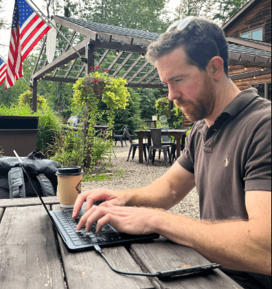 Fotografía de perfil de James O'Beirne sentado en una mesa de madera al aire libre mientras escribe concentrado en una computadora portátil negra. Viste una camisa polo marrón oscuro y lleva gafas de sol sobre la cabeza. A su lado hay un vaso de café para llevar y una mochila negra. Al fondo se aprecia un patio rústico rodeado de vegetación, una pérgola de madera y banderas de Estados Unidos colgadas.
