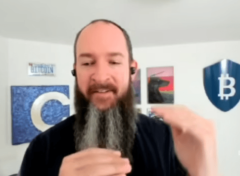 Captura de pantalla de una videollamada que muestra a Jameson Lopp, un hombre con barba larga trenzada o canosa y camiseta negra, gesticulando con las manos mientras habla. De fondo se observa una pared blanca con elementos decorativos temáticos de Bitcoin, incluyendo una placa con la palabra "BITCOIN", un cuadro con la letra "C" azul y un escudo azul con el símbolo de Bitcoin.