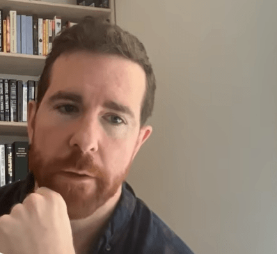 Primer plano de James O'Beirne, un hombre con barba corta rojiza y camisa azul, apoyando la barbilla sobre la mano mientras escucha con gesto pensativo y serio durante una videollamada. Al fondo se observa una biblioteca con libros organizados y una pared clara.