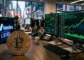 Moneda física de bitcoin con gráficas verdes en el fondo y el logo de Nasdaq.