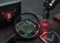 Lupa enfocando un microchip con láser y caja con logo de Bull Bitcoin, representando inspección.