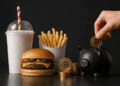 Hamburguesa y papas junto a monedas de Bitcoin y una alcancía que representa el ahorro en criptomonedas.
