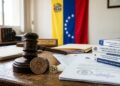 Moneda física de bitcoin con un mazo judicial, documentos y leyes, con la bandera de Venezuela en el fondo.