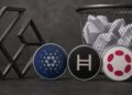Monedas físicas con los logos de Cardano, Hedera y Polkadot junto a una papelera de reciclaje.