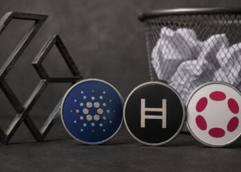 Monedas físicas con los logos de Cardano, Hedera y Polkadot junto a una papelera de reciclaje.