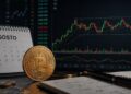 Una moneda física de bitcoin delante de un calendario y un gráfico de precio