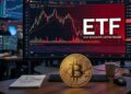 Moneda física de bitcoin dorada sobre una mesa con un monitor que muestra gráficas rojas en el fondo y la palabra ETF.