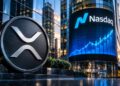 Moneda plateada con el logo de XRP en primer plano frente a una pantalla del edificio Nasdaq.