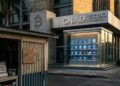 Fotografía editorial en proporción 2:1 de un edificio institucional moderno con el logo de Bitcoin y la sigla CNAD en la fachada. En la entrada, una vitrina de cristal exhibe fichas y tokens digitales brillantes sin utilizar. En primer plano, la fachada de un pequeño comercio local salvadoreño con un cartel que indica cobro en dólares en efectivo.