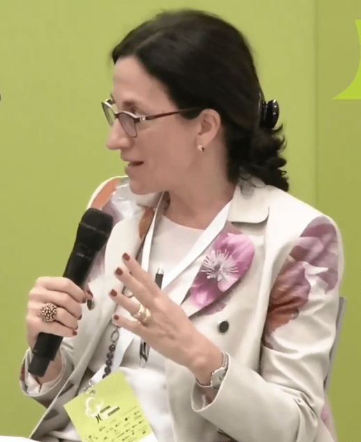 Elena Carletti, mujer de cabello oscuro recogido, lentes de marco fino y blazer crema con estampado floral, habla de perfil sosteniendo un micrófono con la mano derecha mientras gesticula con la izquierda en un panel institucional.