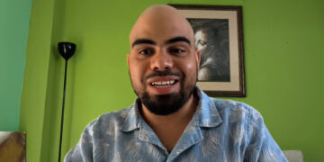 Forte 11, fundador de Cuba Bitcoin y el entrevistado del podcast Separando el dinero del Estado de criptoNoticias. Forte11 está sonriendo con camisa azul estampada sobre un fondo de pared verde.