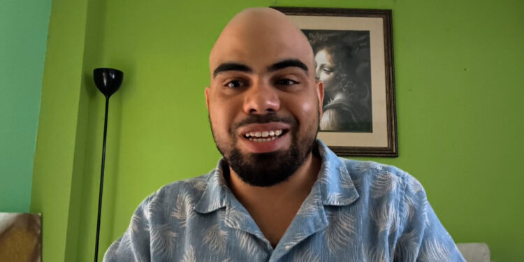 Forte 11, fundador de Cuba Bitcoin y el entrevistado del podcast Separando el dinero del Estado de criptoNoticias. Forte11 está sonriendo con camisa azul estampada sobre un fondo de pared verde.