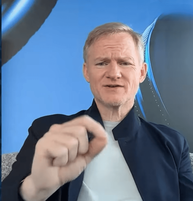 Fotograma en primer plano de Mike Belshe, CEO de BitGo, con el pelo gris, chaqueta azul oscuro y camiseta gris. Apunta con el dedo índice directamente hacia la cámara con una expresión seria y enfática. El fondo es un azul brillante con líneas curvas abstractas y un logotipo parcialmente visible en la esquina superior derecha.