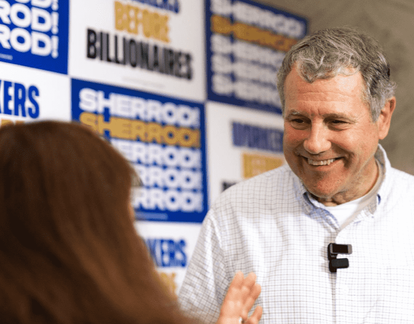 El político estadounidense Sherrod Brown sonríe mientras conversa con una mujer en primer plano de espaldas. Viste una camisa a cuadros clara con un micrófono de solapa. De fondo se observan carteles publicitarios azulez y blancos con su nombre y la consigna "Workers Before Billionaires".