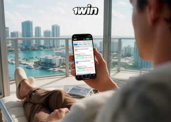 un hombre, sentado frente a ventanales con una ciudad costera de fondo, sosteniendo un celular que muestra la plataforma de trading de Win.