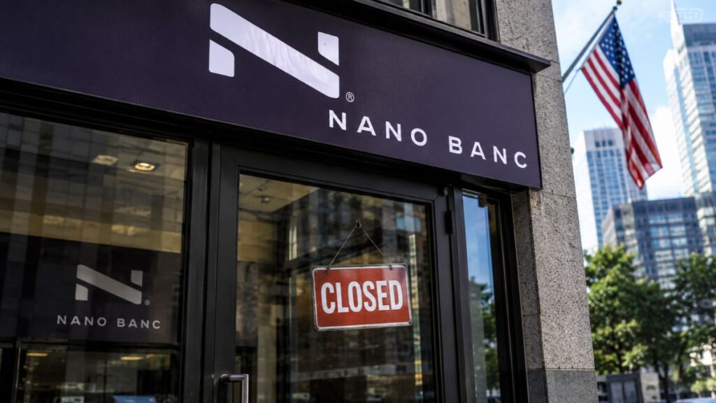 Fachada de una sucursal bancaria de Nano Banc con un letrero de cerrado en la puerta y la bandera de Estados Unidos al fondo.