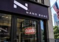 Fachada de una sucursal bancaria de Nano Banc con un letrero de cerrado en la puerta y la bandera de Estados Unidos al fondo.