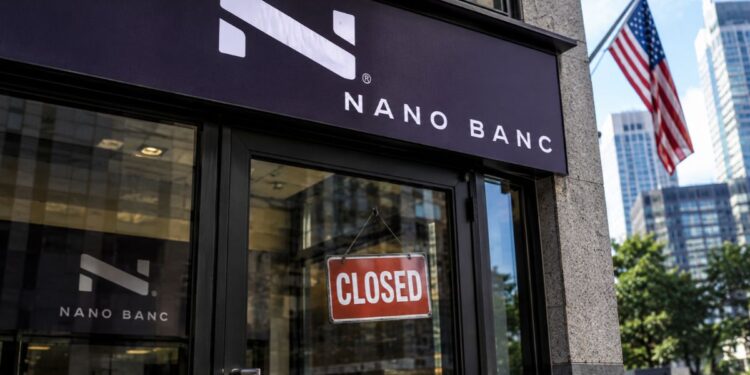 Fachada de una sucursal bancaria de Nano Banc con un letrero de cerrado en la puerta y la bandera de Estados Unidos al fondo.