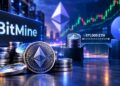 Una pila de criptomonedas Ethereum en primer plano con un gran monitor en el fondo que muestra el logotipo de BitMine.