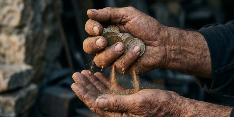 Primer plano de unas manos trabajadoras —de alguien mayor, o con marcas de oficio— sosteniendo o guardando unas monedas, mientras estas parecen desvanecerse o erosionarse en polvo entre los dedos. Luz cálida y humana sobre las manos, fondo neutro y frío. Contrasta el trabajo guardado (tiempo humano) con la dilución silenciosa. Paleta cálida en el sujeto, fría en el entorno, subrayando el axioma del "trabajo guardado".