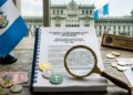 un escritorio de madera con un libro legal, una lupa enfocando el documento, monedas de criptomonedas (Bitcoin y Ethereum) y la bandera de Guatemala.