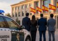 Cuatro personas trajeadas de espaldas frente a un edificio oficial con banderas de España y un coche de la Guardia Civil.