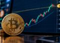 Moneda física de Bitcoin dorada junto a una pantalla con un gráfico financiero alcista alcanzando los USD 85.000.