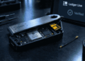 Hardware wallet abierta con un módulo espía amarillo oculto bajo la carcasa, mientras una pantalla al fondo muestra la verificación de Ledger Live como genuina.