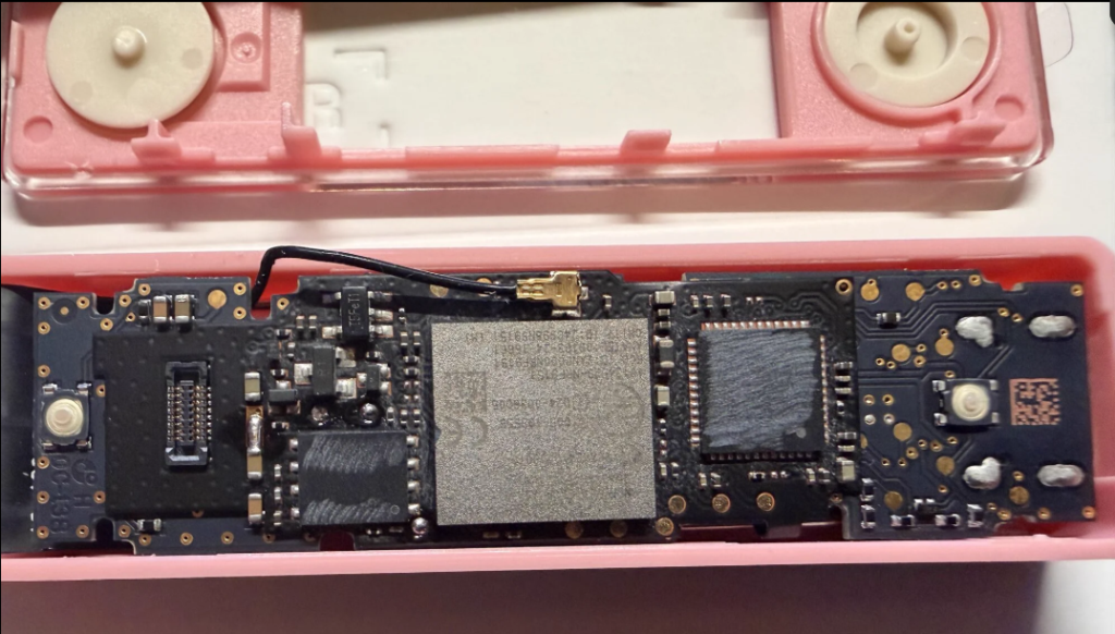 Placa base de una hardware wallet desmontada dentro de una carcasa de plástico rosa. Se observa la carcasa superior con dos botones circulares en la parte de arriba. En la placa inferior, se aprecian dos blindajes metálicos que cubren los chips principales y el elemento seguro, junto a un cable de antena negro conectado a un puerto U.FL en el centro de la placa. La placa está expuesta sobre la bandeja rosa.