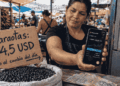 Fotografía de un puesto de mercado en Venezuela. Un cartel de cartón escrito a mano dice "Caraotas: 45 USDT" con una flecha que indica "o al cambio del día". El vendedor sostiene un teléfono inteligente con la aplicación Binance P2P mostrando la cotización del USDT. En la pared hay un código QR pegado con cinta adhesiva. Al fondo se ven otros puestos y clientes comprando.
