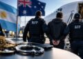 Agentes de la AFP escoltan a un detenidopospos en un aeropuerto hacia un avión, junto a banderas de Argentina y Australia y monedas de Bitcoin.